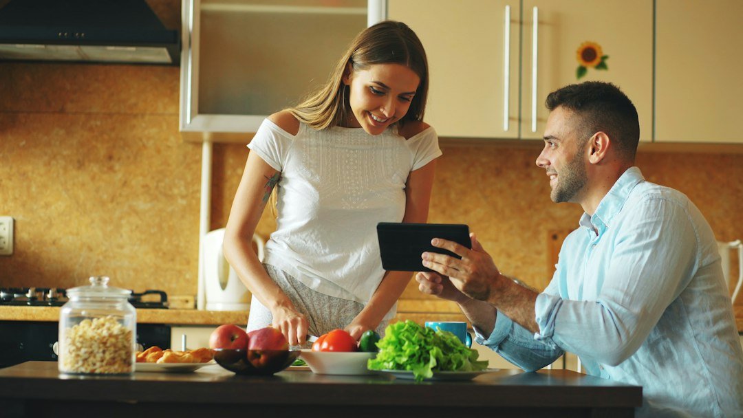 🎬 FULL 4K VIDEO for this still is in my bio link! ⬇️ Attractive couple chatting in the kitchen early morning. Handsome man using tablet while his girlfriend cooking breakfast at home