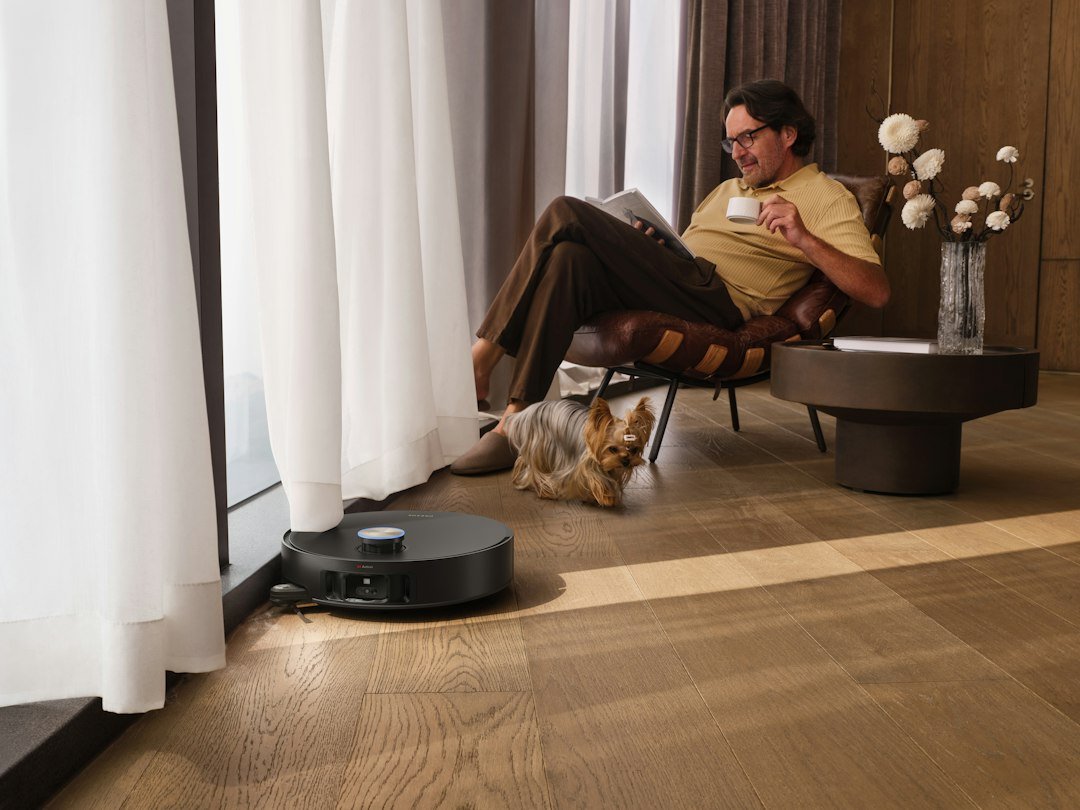 A man relaxes comfortably in a stylish armchair by a sunlit window, reading a book and enjoying a warm drink, while a robot vacuum cleaner quietly keeps the hardwood floor clean beside him.
